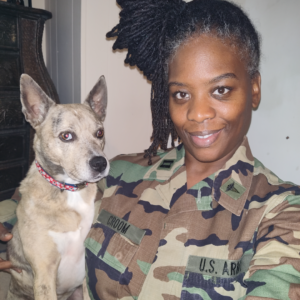 Black female army veterinary officer in army fatigues sitting with her tan brindle cattle dog.